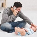 A man talking on the phone while holding a baby on a bed.