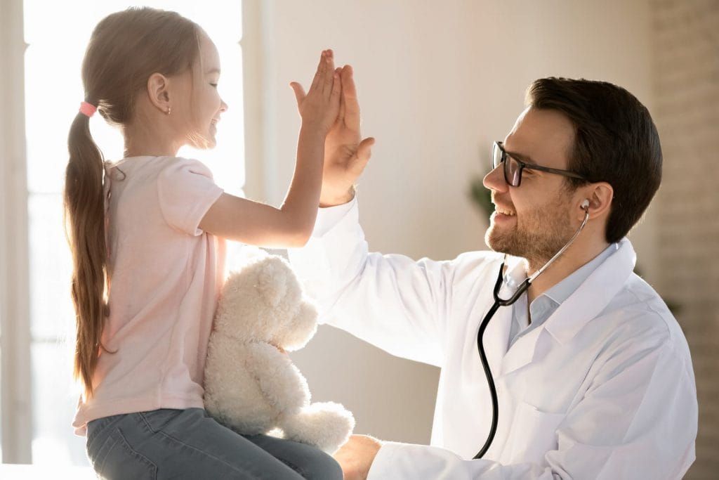 A smiling pediatrician giving a high-five to a young girl holding a teddy bear in Bellaire, TX.