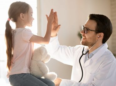 A smiling pediatrician giving a high-five to a young girl holding a teddy bear in Bellaire, TX.